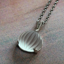 Load image into Gallery viewer, A silver-toned pendant necklace featuring a seashell-shaped pendant with a textured, ribbed surface, resting on a textured, mottled surface.
